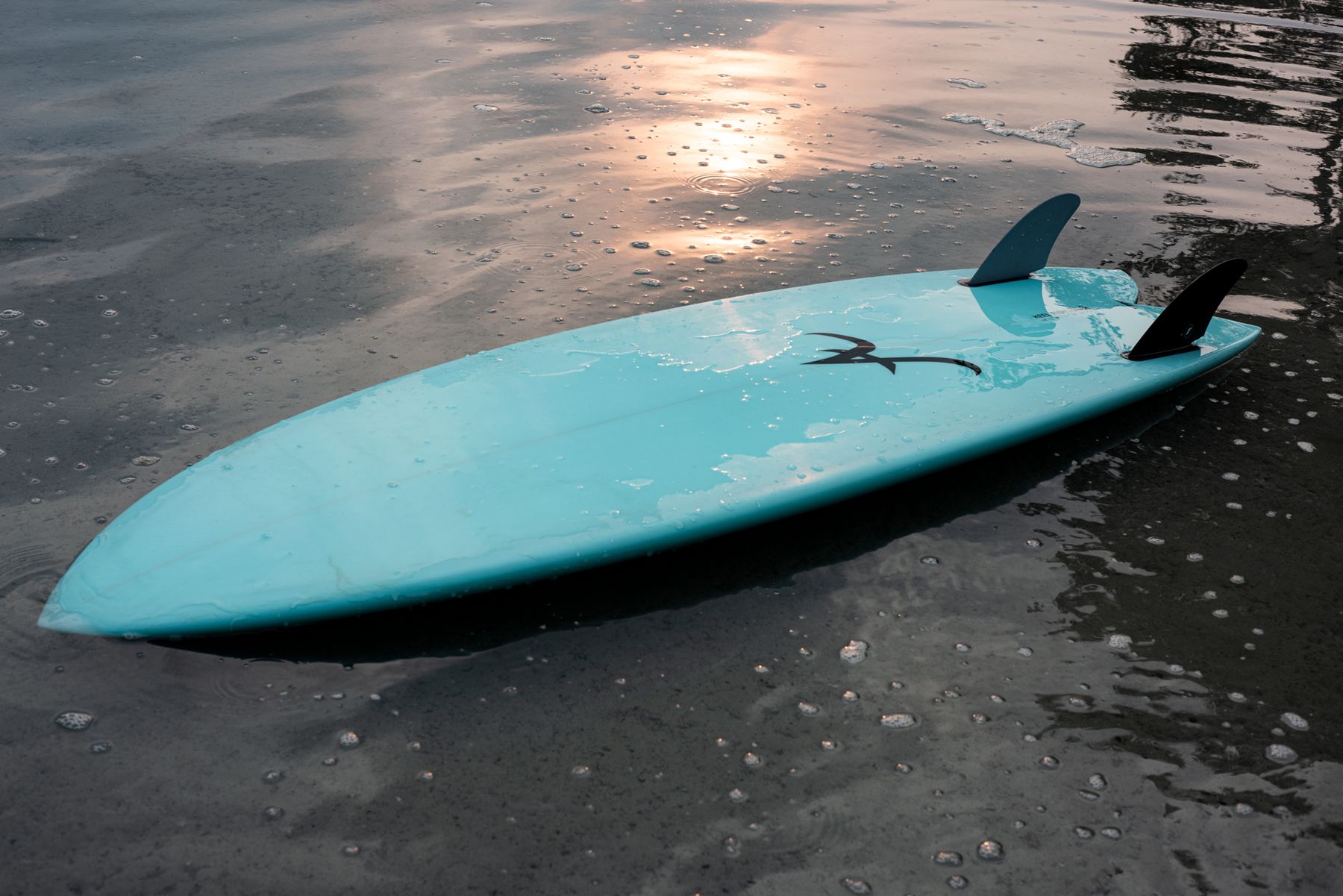 An Aloha twin-fin surfboard resting in shallow water at sunset
